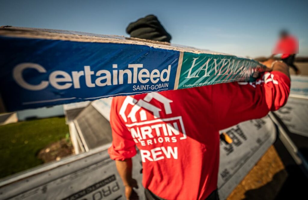 Martin Exteriors roofer carrying a bundle of shingles across a roof deck covered in underlayment