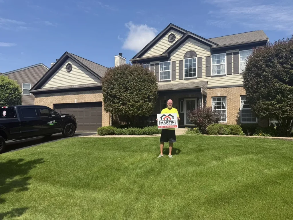 Smiling Martin Exteriors Roofing & Siding customer holding a yard sign on the front lawn of their two-story home