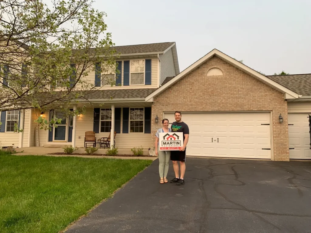 Smiling Martin Exteriors Roofing & Siding customers holding a yard sign in the driveway of their brick two-story home