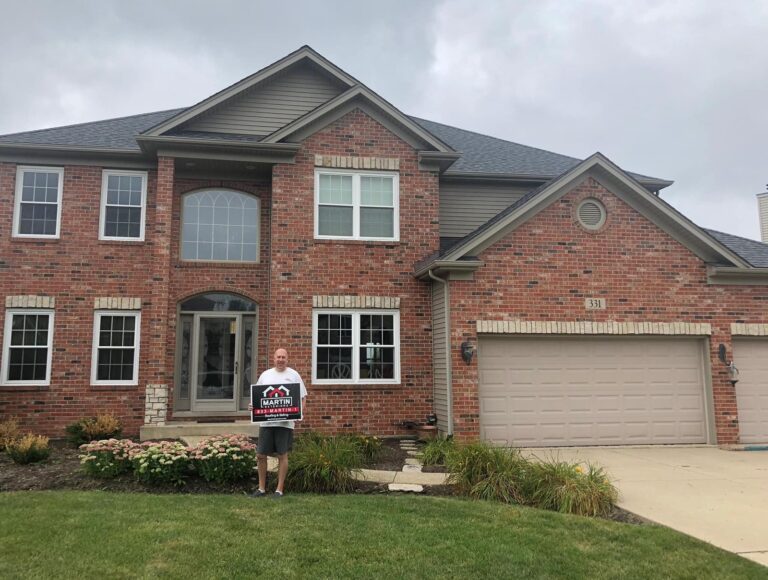 Martin Exteriors project in Rockford, IL: homeowner holding a Martin sign in front of a two-story brick home with a gray shingle roof
