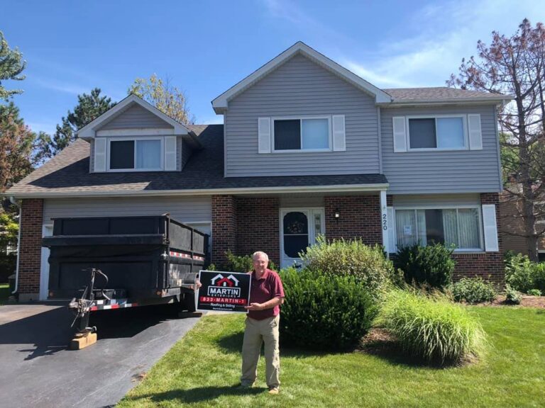 Martin Exteriors project in Rockford, IL: homeowner holding a Martin sign in front of a two-story home with gray vinyl siding and brick, with a Martin dump trailer in the drive